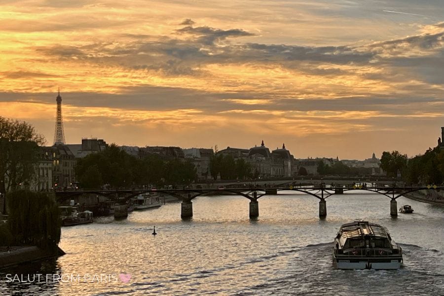 A golden sunset over the Seine in Paris in May with the Eiffel Tower in the distance, a boat moving along the river, and a bridge silhouetted against the glowing sky.