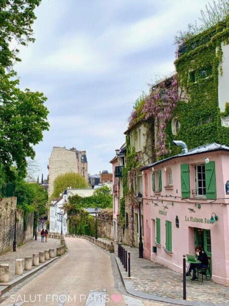 A quiet cobblestone street in Paris in May lined with ivy covered buildings and a pink cafรฉ with green shutters, where a person sits outside and wisteria blooms overhead.