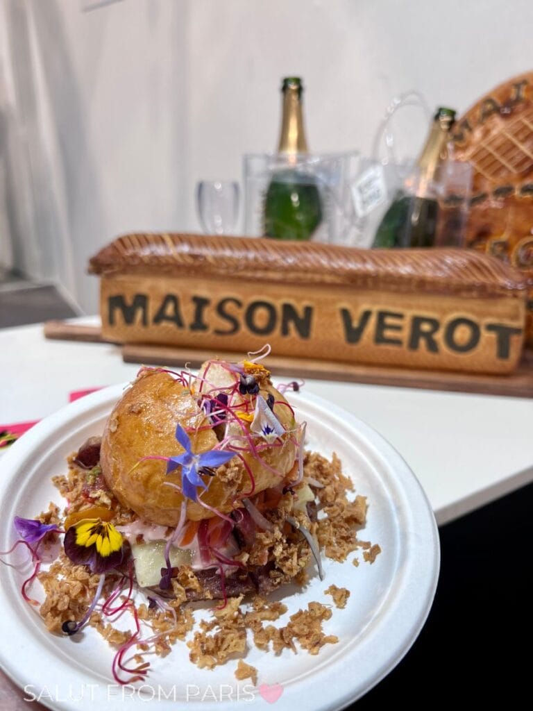 A plated dish topped with edible flowers and crispy garnish sits in front of a sign reading "MAISON VEROT," with champagne bottles in the background on a table.