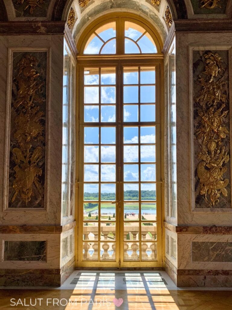 Sunlight streams through tall ornate windows inside a grand palace in Paris in July, revealing gilded details and a view of manicured gardens and water beyond.